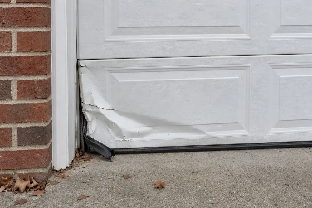 Crumpled bottom corner of a white raised-panel garage door with cracked paint and a torn bottom seal